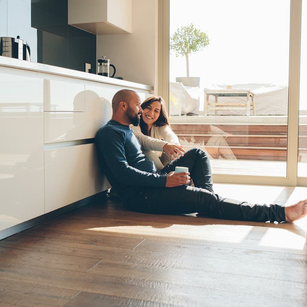 Ein Paar sitzt am Boden an einer Küchenzeile und trinkt Kaffee im Sonnenlicht.
